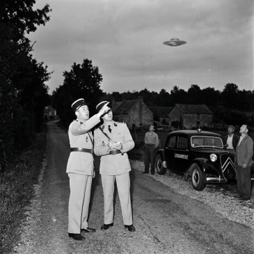 Two French gendarmes in uniform pointing at a UFO in the sky with villagers and a police car nearby