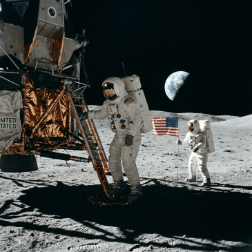 Two astronauts in white spacesuits on the moon next to a lunar module and American flag with Earth in background