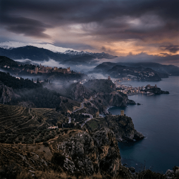 Coastal town with cliffs, mist, and snowy mountains at sunset